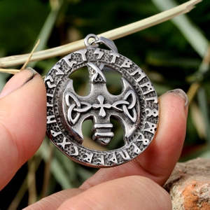 Close-up of a rustic silver cross pendant with runic carvings held by hand in nature