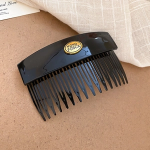 Detail of a black acrylic hair comb featuring a gold oval plaque with engraved letters