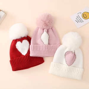 Knitted infant hats with heart patterns and faux fur pom-poms in red, pink and white