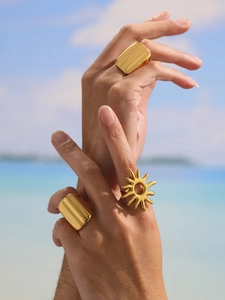 Close-up of golden sunburst and wide chunky metal rings on hand with beach background