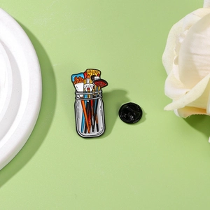 Artistic enamel pin featuring colorful paint brushes in a glass jar on a soft green background