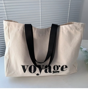 Detail of a large off-white canvas tote bag featuring black voyage lettering and handles