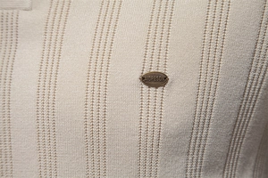 Detailansicht der hellbeigen Strickstruktur mit vertikalen Streifen und einem Metallschild