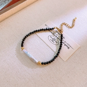 Detail of a thin black beaded bracelet featuring a long white baroque pearl and gold clasp