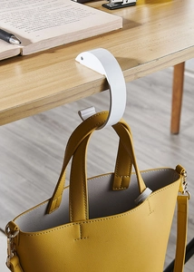 Close-up of white portable desk hook hanging a yellow bag on a wooden table edge