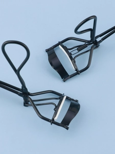 Close-up of ergonomic black eyelash curlers with white silicone pads on a blue background