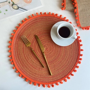 Macro detail of round orange woven placemat with ball fringe and golden cutlery set