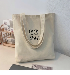 Detalle del bolso tote de lona beige con estampado de ojos negros y el texto shh!