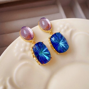 Close-up of geometric gold earrings with purple and gradient blue crystals on a ceramic plate