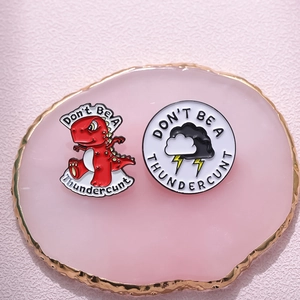 Macro shot of red dinosaur and round enamel pins with witty text on a pink marble slab