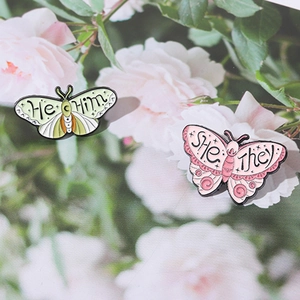 Dettaglio di spille a farfalla smaltate con pronomi He/Him e She/They su fiori