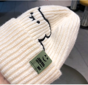 Close-up of beige knit beanie showing ribbed texture, cartoon embroidery and green woven label