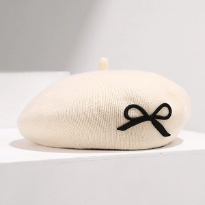 Détail d'un béret en maille blanc cassé avec un nœud noir sur une plateforme épurée