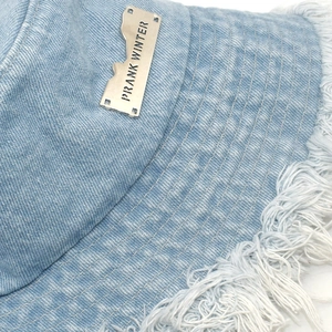 Macro detail of frayed denim edge and silver metal logo tag on light blue bucket hat