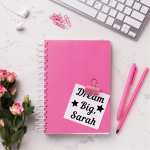 Flat lay of pink spiral notebook with custom labels, pens and stationery on white desk