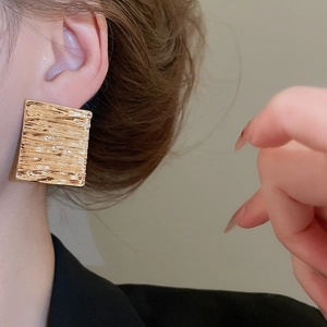 Detail shot of large square gold earrings with brushed metallic texture worn by a model
