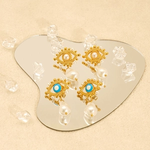 Gold evil eye earrings with blue enamel and pearl drops on a sandy texture background