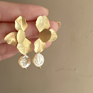 Detail showing golden flower petal earrings with baroque pearls held in hand for scale