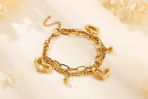 Détail macro du bracelet multicouche en acier doré avec breloques cœur et perles rondes