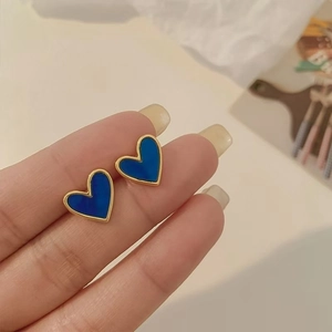 Macro shot of blue heart stud earrings with gold trim held on fingertips for scale