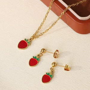 Close-up of golden necklace and earrings with red enamel strawberry charms on beige fabric