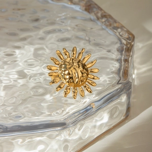 Détail d'une bague soleil dorée avec texture martelée sur un plateau en verre ondulé