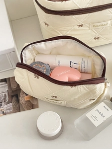 Dettaglio del ricamo a fiocco trapuntato sulla borsa cosmetica color crema con cerniera