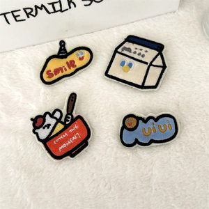 Detail of four cartoon embroidered brooches featuring a smiley face and milk box on plush background