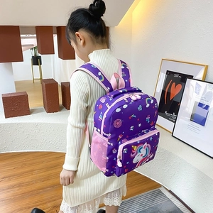 Child wearing a purple cartoon unicorn backpack with pink mesh side pockets in a bright room