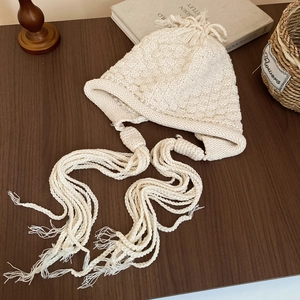 Detailed close-up of beige chunky knit earflap hat with tassels on a wooden surface