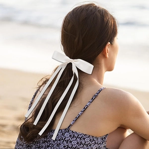 Elegant white satin bow hair clip with long ribbons worn by a woman at the beach