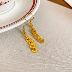 Macro shot of long gold-tone coin-style earrings on beige fabric with ceramic saucer background