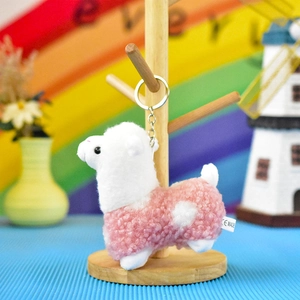 Detail view of a cute pink and white plush alpaca keychain hanging on a wooden stand.