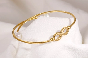 Detail van gouden gedraaide armband met oneindigheidssymbool en witte parels