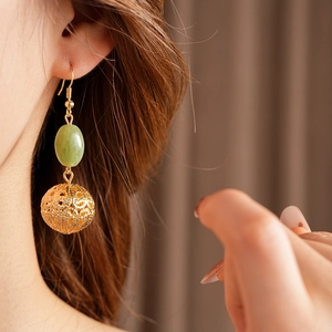 Detail view of gold earring with green gemstone and hollow metal pendant on model