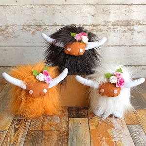 Three highland cow plush toys with floral crowns in black, orange, and white faux fur