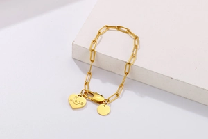 Detailed close-up of golden paperclip chain bracelet with engraved heart and round charms