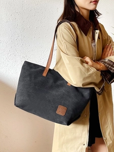 Large black canvas tote bag with brown leather straps worn by a model in a beige coat