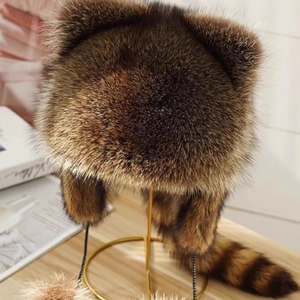 Faux raccoon fur trapper hat with ears and long tail on a gold display stand