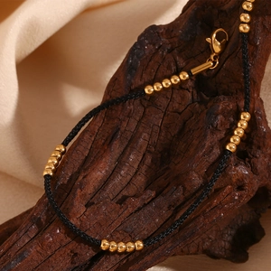 Detailed shot of 18K gold-plated beads on a black cord anklet draped over rustic wood