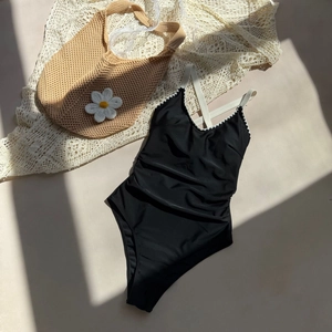 Flat lay of black one-piece swimsuit with white wavy trim and crossover straps on beige background