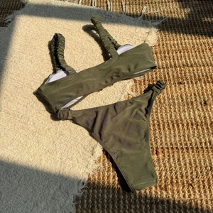Detailed flat lay of olive green ribbed bikini with ruched straps on a woven rug
