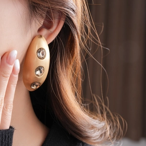 Close-up of golden matte oval earrings with crystals worn by a model in soft light