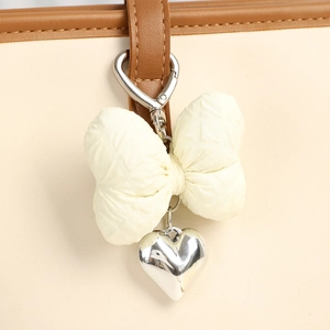 Quilted cream bow keychain with silver heart pendant on a white leather handbag strap