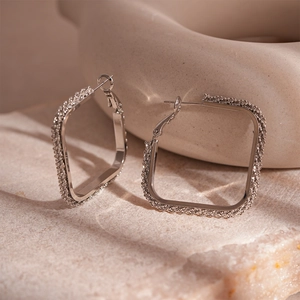 Macro shot of square silver earrings with textured crushed diamond edges on beige stone