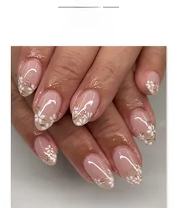 Gros plan d'ongles en amande, base rose nude, motifs floraux blancs et touches dorées