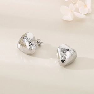 Polished silver heart stud earrings with hammered metal texture on beige background