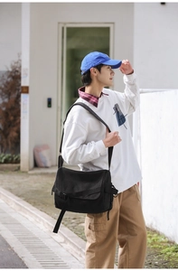 Men's large black canvas messenger bag shown on shoulder in a minimalist outdoor setting