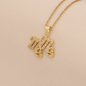 Macro detail of gold-plated MOM cursive pendant with inlaid zircons on beige fabric
