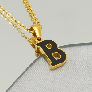Gold-plated stainless steel necklace with a black letter B pendant on reflective surface